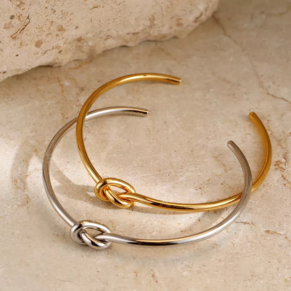 Two intertwining Twist Knot Stainless Steel Cuff Bracelets, one gold and one silver, on a stone surface, embodying new fashion style.