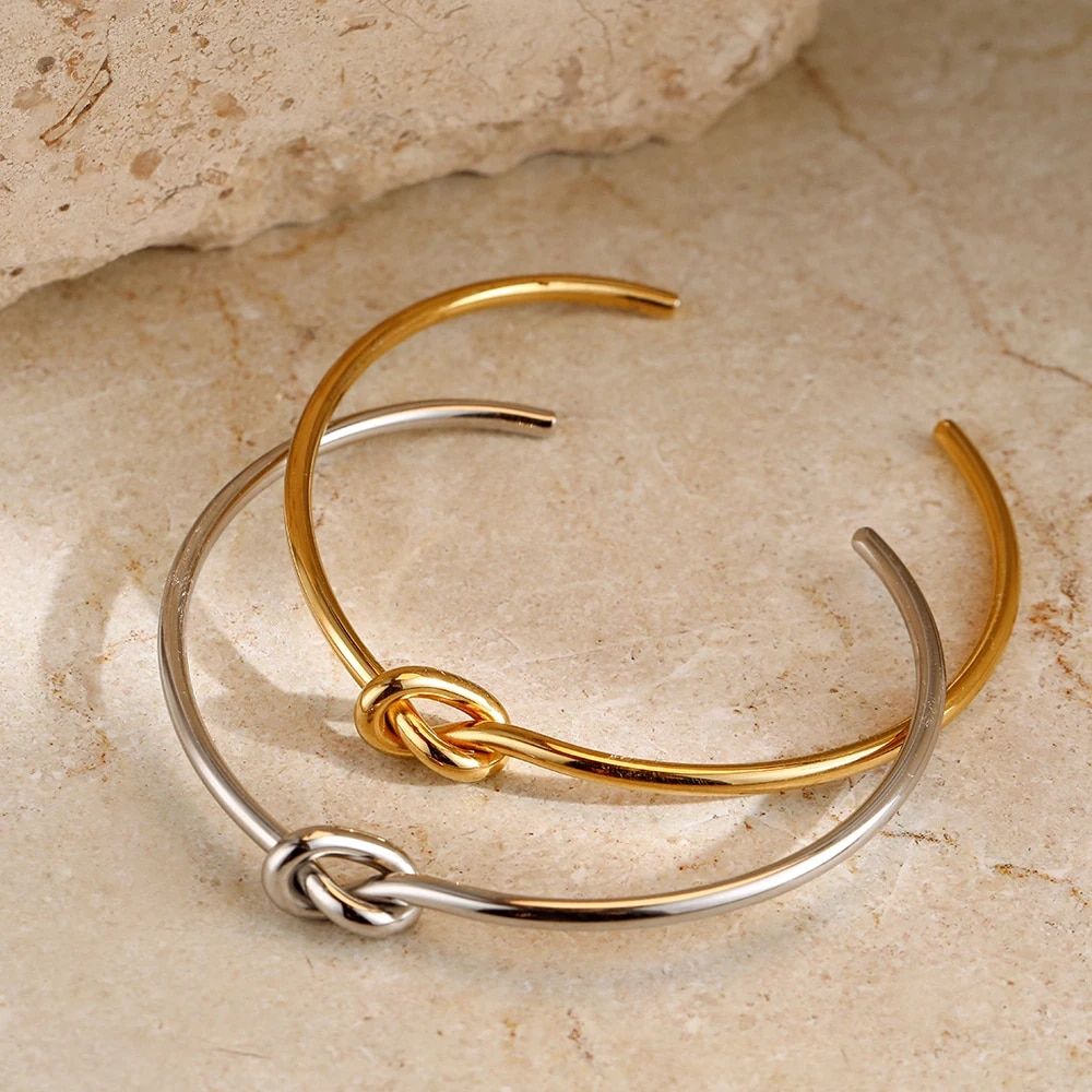 Two intertwined Twist Knot Stainless Steel Cuff Bracelets, one gold and one silver, positioned on a stone surface, represent the latest in fashion accessory trends.