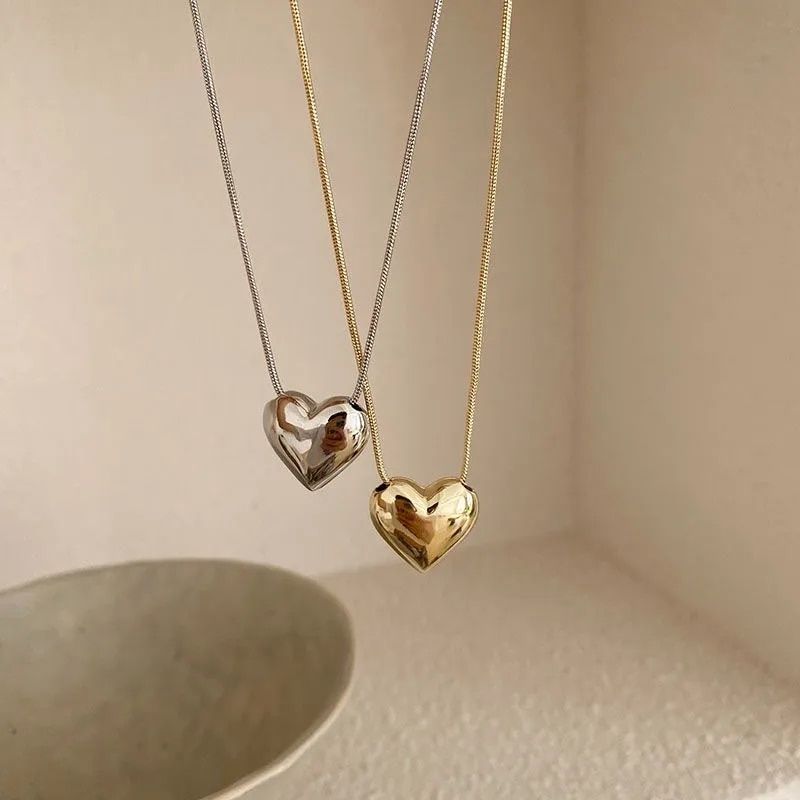 Two Gothic Trendy Heart Pendant Necklaces, one silver and one gold, hanging against a cream background with soft shadows, epitomizing current fashion trends.