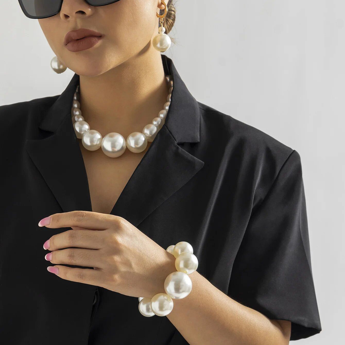 A woman in sunglasses and a black shirt adorned with a Womens Round Pearl Charm Bracelet and necklace, touching her chin thoughtfully.