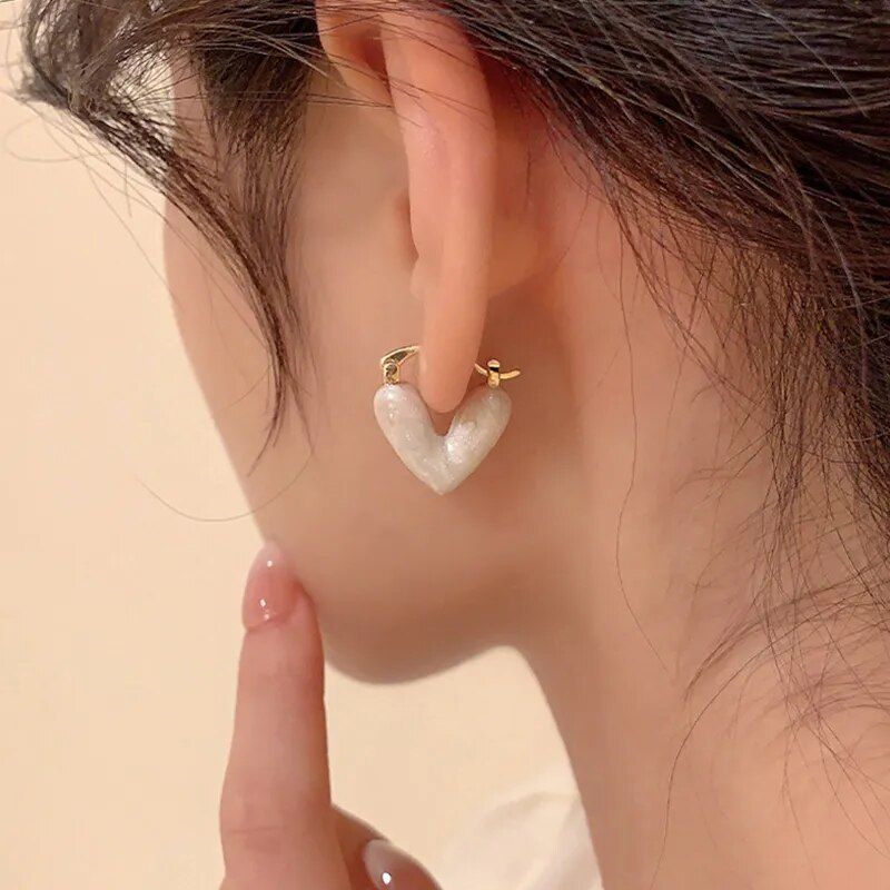 Close-up of a person's ear adorned with Elegant Heart Small Drop Earrings for Women featuring a white heart-shaped pendant, highlighting this fashion accessory with a finger pointing at it.