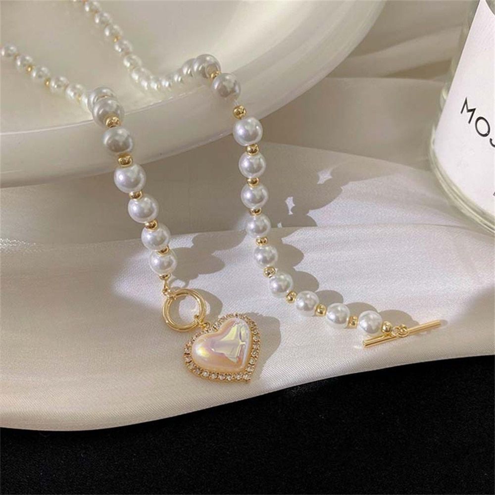 A Vintage Heart Pendant Pearl Necklace with a heart-shaped pendant and gold accents displayed elegantly on a creamy surface, epitomizing current womens fashion trends.