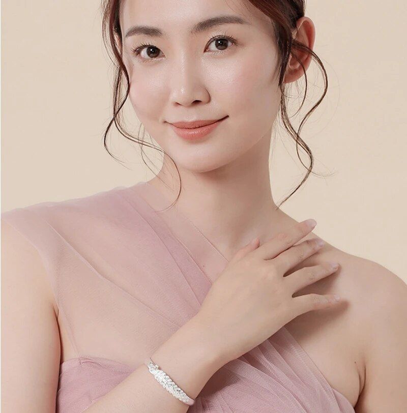 A woman with a gentle smile, wearing a pink dress and a Womens Vintage Peacock Cuff Bracelet - Antique Silver Plated, touches her collarbone lightly, against a peach background. The bracelet is not just an ornament but also a significant fashion accessory