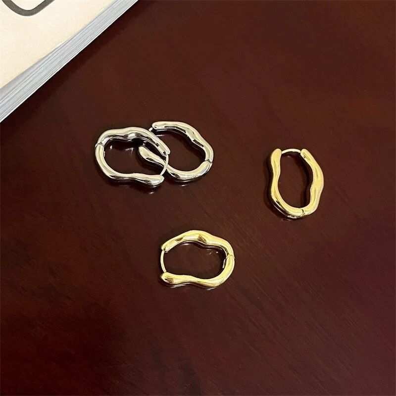 Three Luxurious Geometric Hoop Earrings for Women on a wooden table, inspired by new fashion trends, featuring two silver pieces and one gold piece, resembling irregular loops.