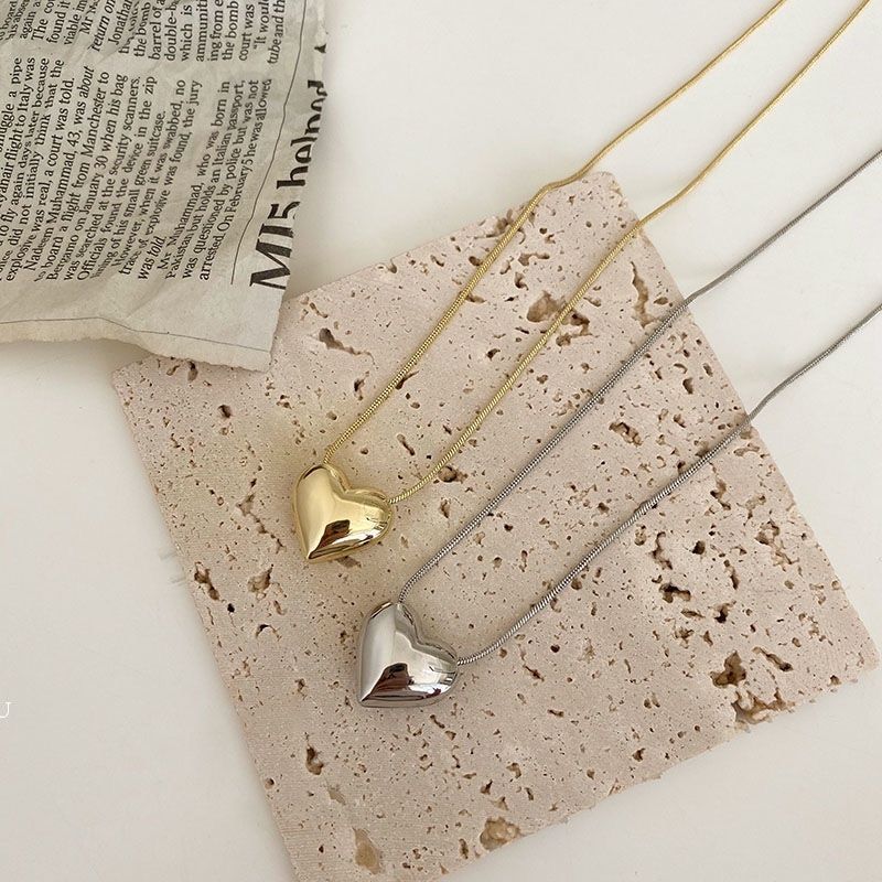 Two Gothic Trendy Heart pendant necklaces, one gold and one silver, representing the latest fashion trends, resting on a textured beige surface beside a folded newspaper.