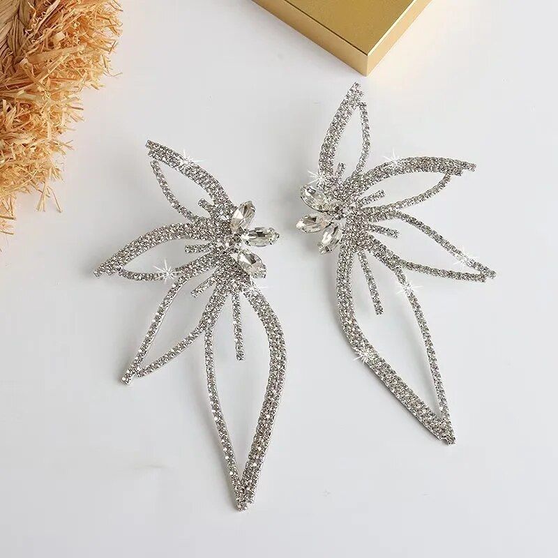 Two large, star-shaped Chic Maple Leaf Zircon Earrings adorned with multiple small crystals, displayed on a light beige surface next to a dried bouquet and a gift box. These earrings are the latest in new fashion trends.