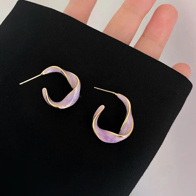 A pair of Lavender Purple Twisted Hoop Earrings, a trendy fashion accessory with a purple iridescent finish, displayed on a black background held by fingers.