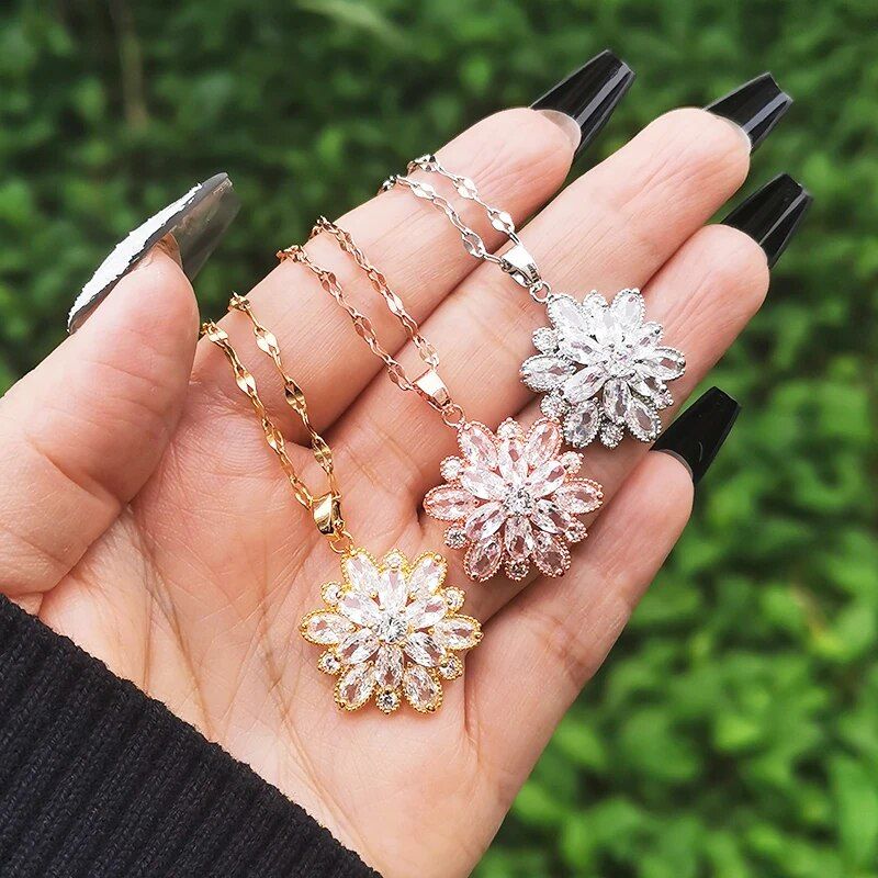 A person's hands holding four Womens Gold-Plated Crystal Flower Pendant Chokers against a green leafy background, epitomizing current fashion trends.