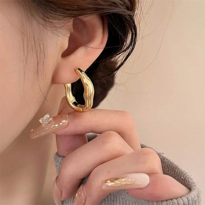 A woman with luxurious geometric hoop earrings adjusts her clear, floral acrylic nail near her ear, showcasing new fashion jewelry and nail art.