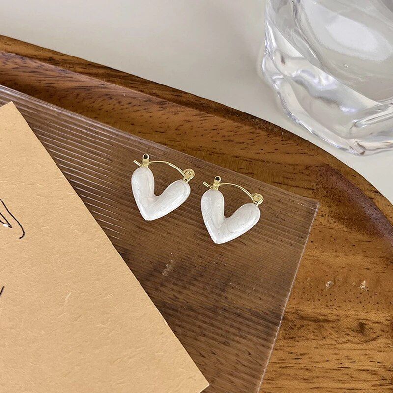 A pair of Elegant Heart Small Drop Earrings for Women, a trendy fashion accessory, placed on a clear display stand with a piece of brown textured paper and a glass cup in the background.