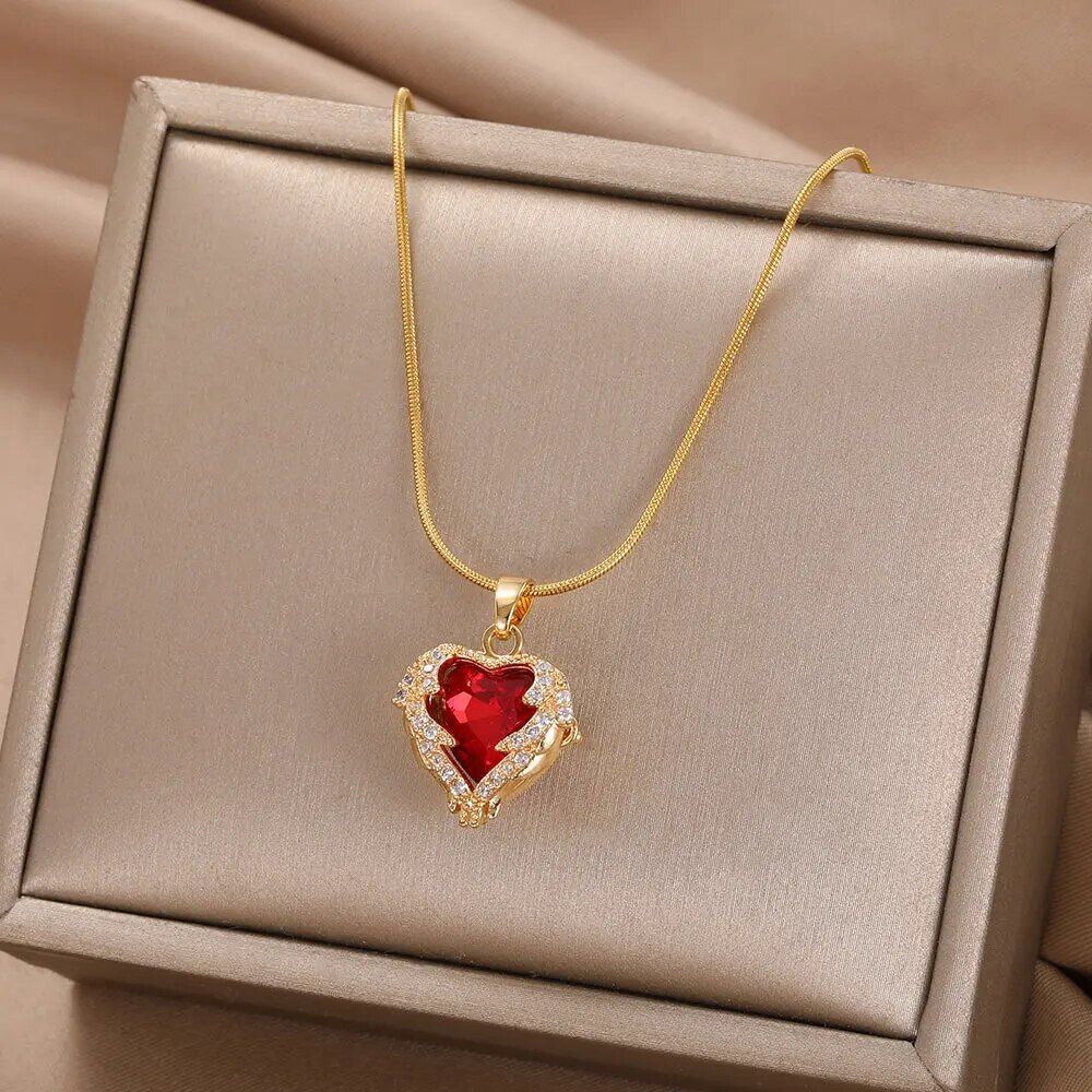 A Gold-Plated Enamel Red Heart Pendant Necklace with a large red gemstone and small diamonds, displayed in an open beige box, embodies the latest fashion trends.