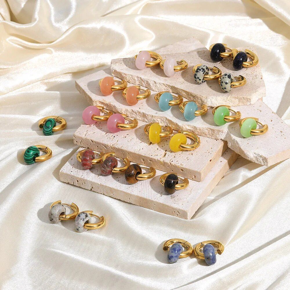 A collection of colorful gemstone rings displayed on textured beige stone slabs, set against a silky cream fabric backdrop, ideal for complementing Women's Natural Stone Hoop Earrings.